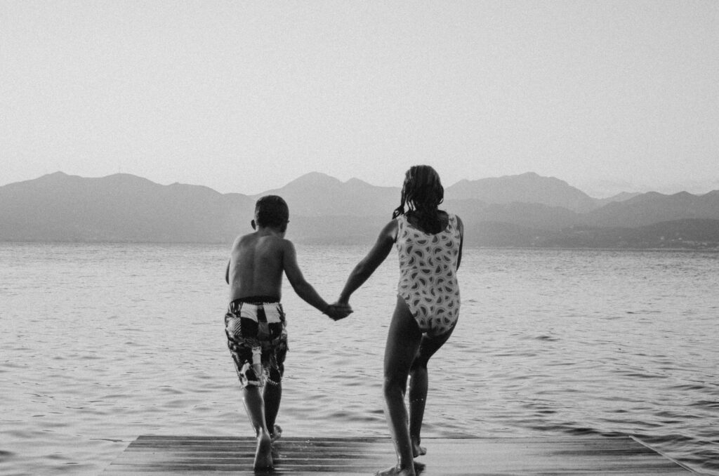 grayscale photography of girl and boy standing beside body of water